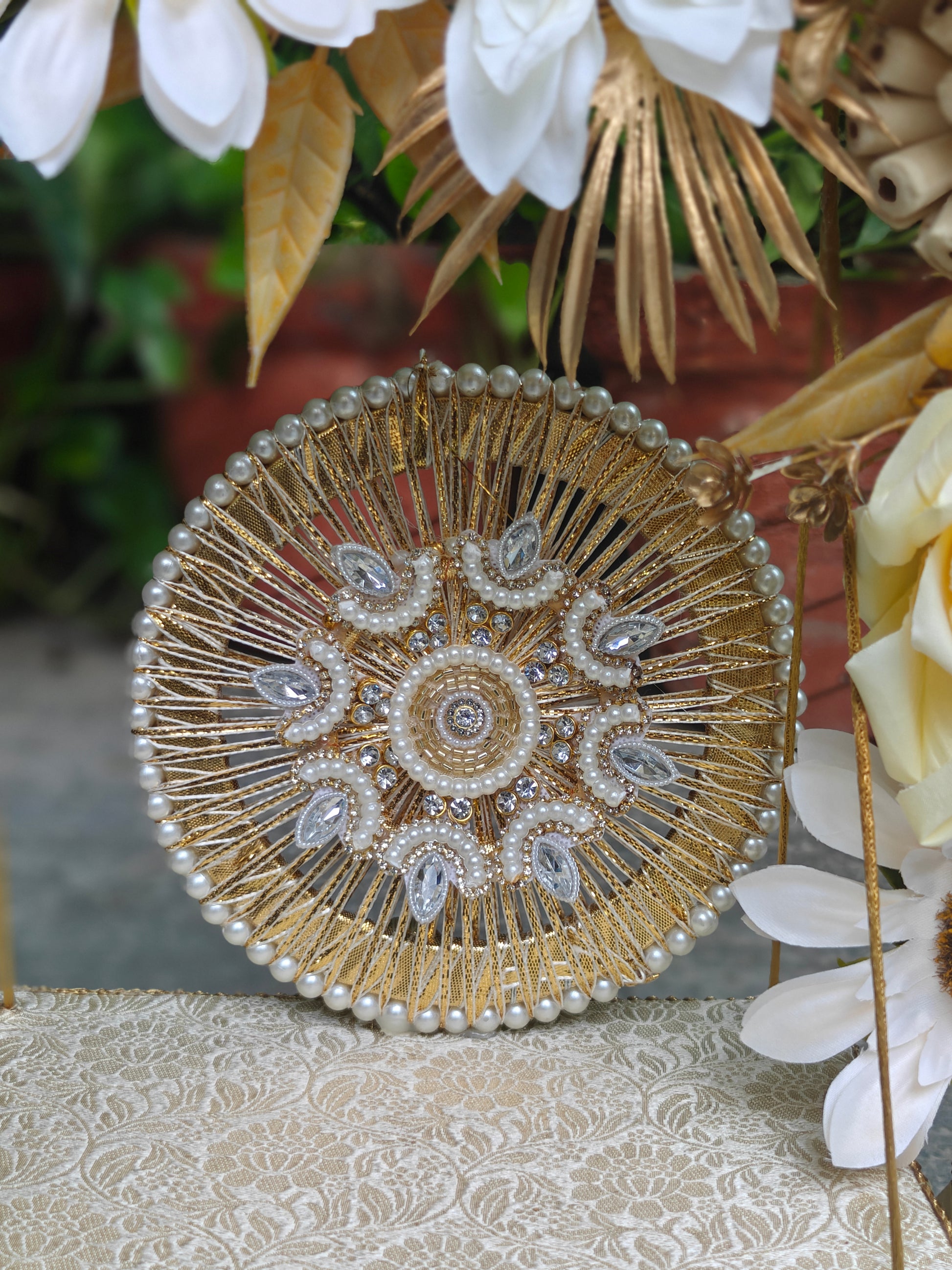A round gold beaded and embroidered decorative piece with pearl-like beads and clear rhinestone accents, displayed on a patterned cream fabric with white flowers and gold leaves in the background.