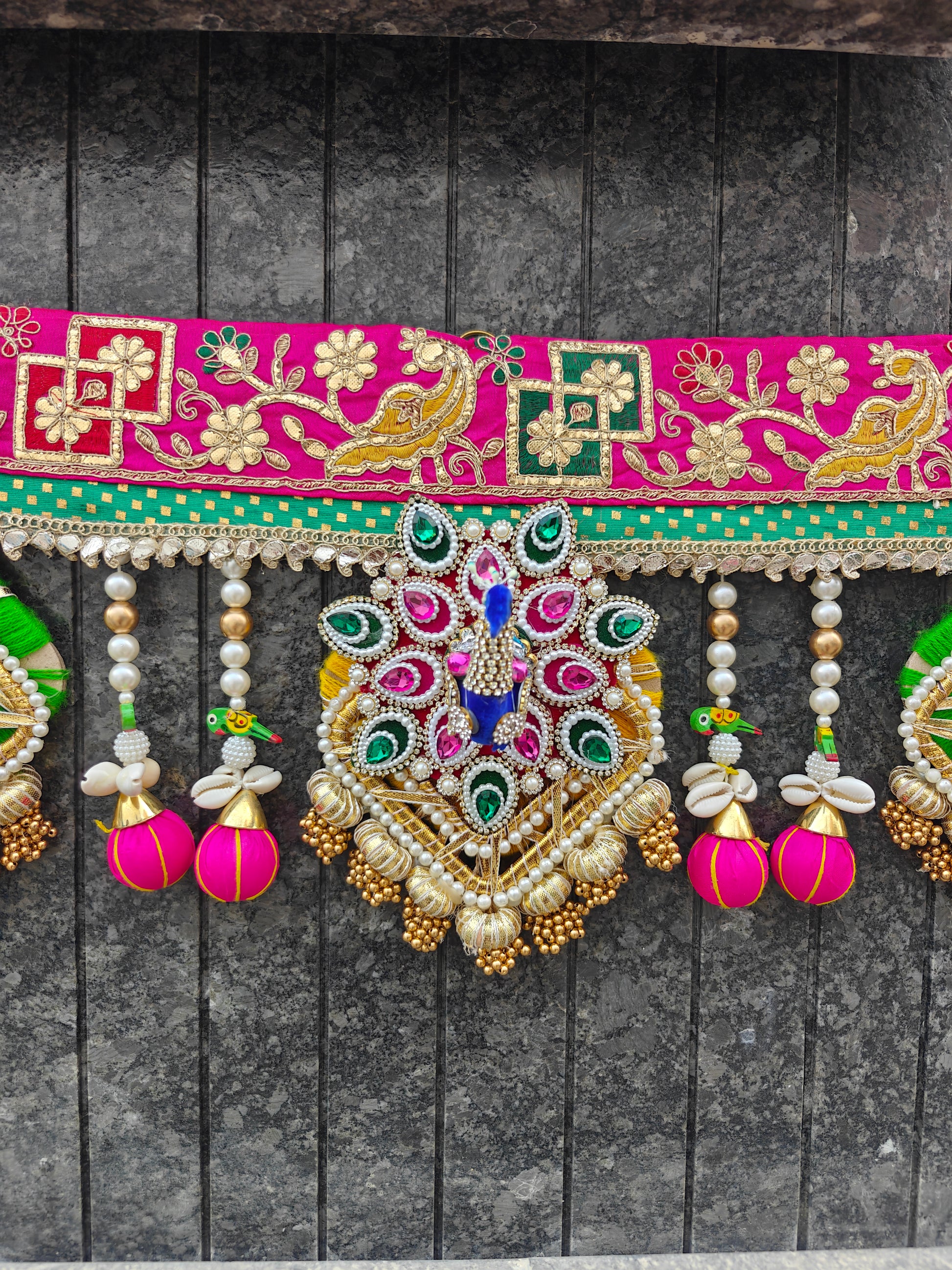 A close-up of a vibrant traditional Indian decorative garland or sari panel hanging against a dark gray paneled wall. The fabric is bright pink with intricate gold embroidery, green accents, and a large central peacock motif embellished with rhinestones in blue, green, pink, and gold.