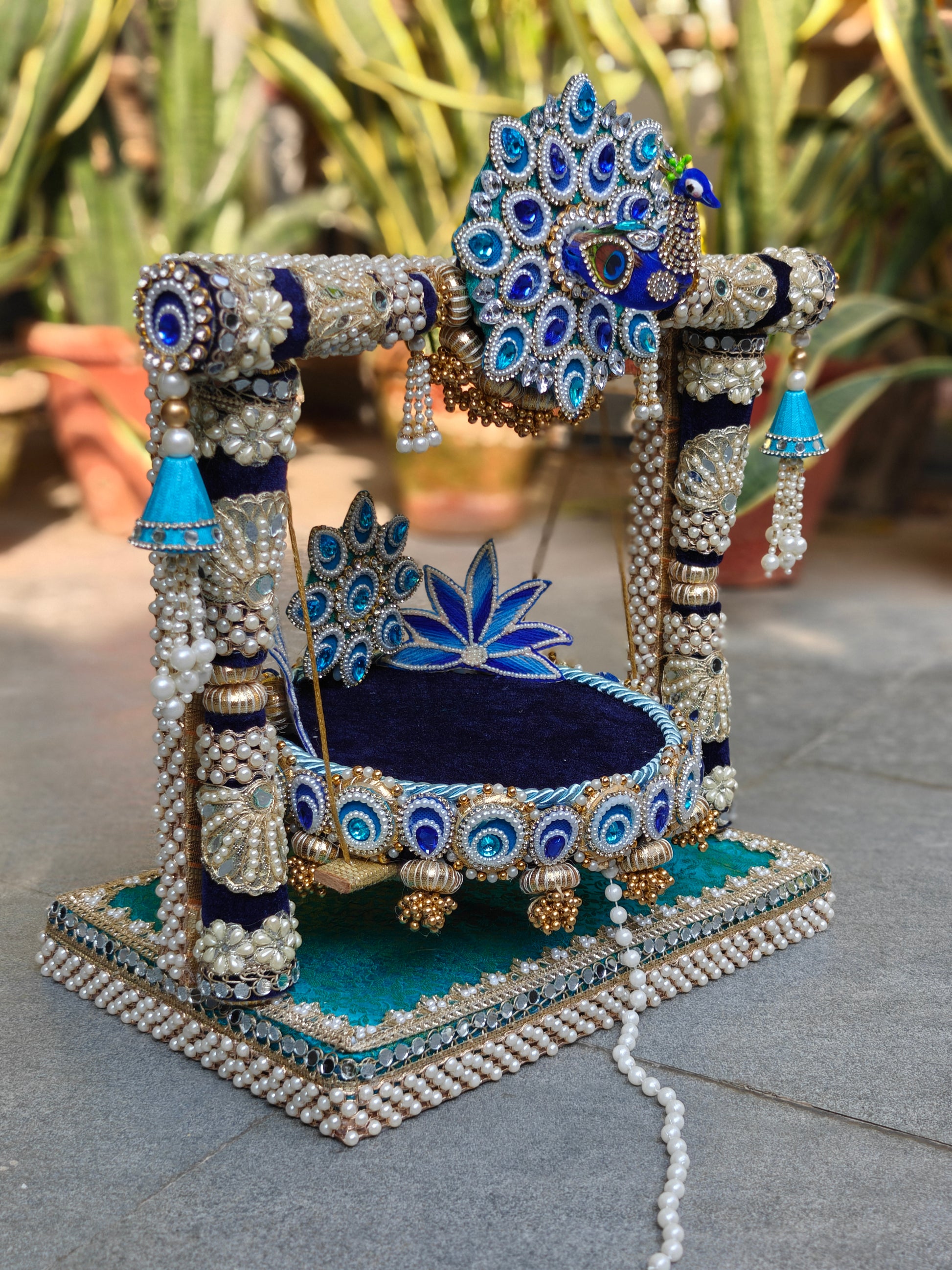 Side view of Royal Collection 2-in-1 handcrafted Ladoo Gopal peacock jhula in royal navy blue and teal velvet – 3D enamel peacock torana crown with sapphire and aqua teardrop stones, navy and silver zardozi pillar frame with pearl clusters, sky blue ghungroo corner bells, detachable navy patra with peacock lotus backrest and gold ghungroo legs, teal MDF base with double pearl border – The Wedding Station