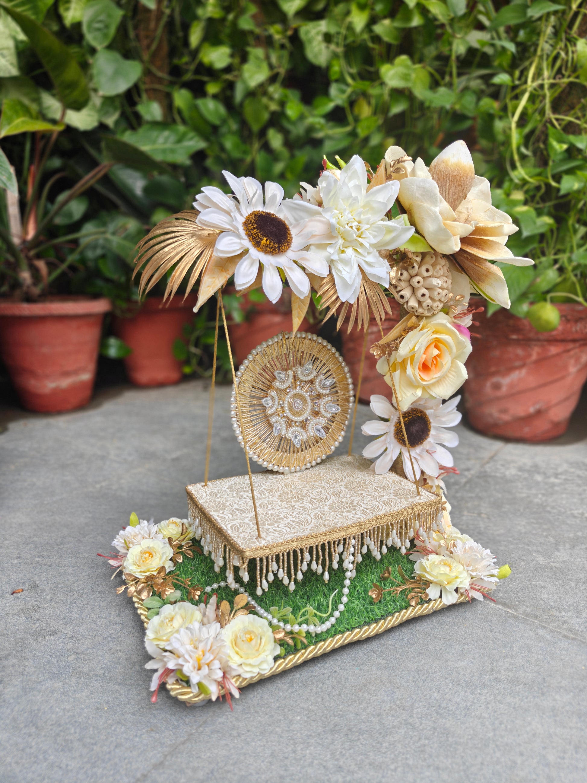 A decorative handmade floral display stand with faux flowers, a woven round ornament, and a lace-covered platform, photographed outdoors against potted plants on a stone surface.