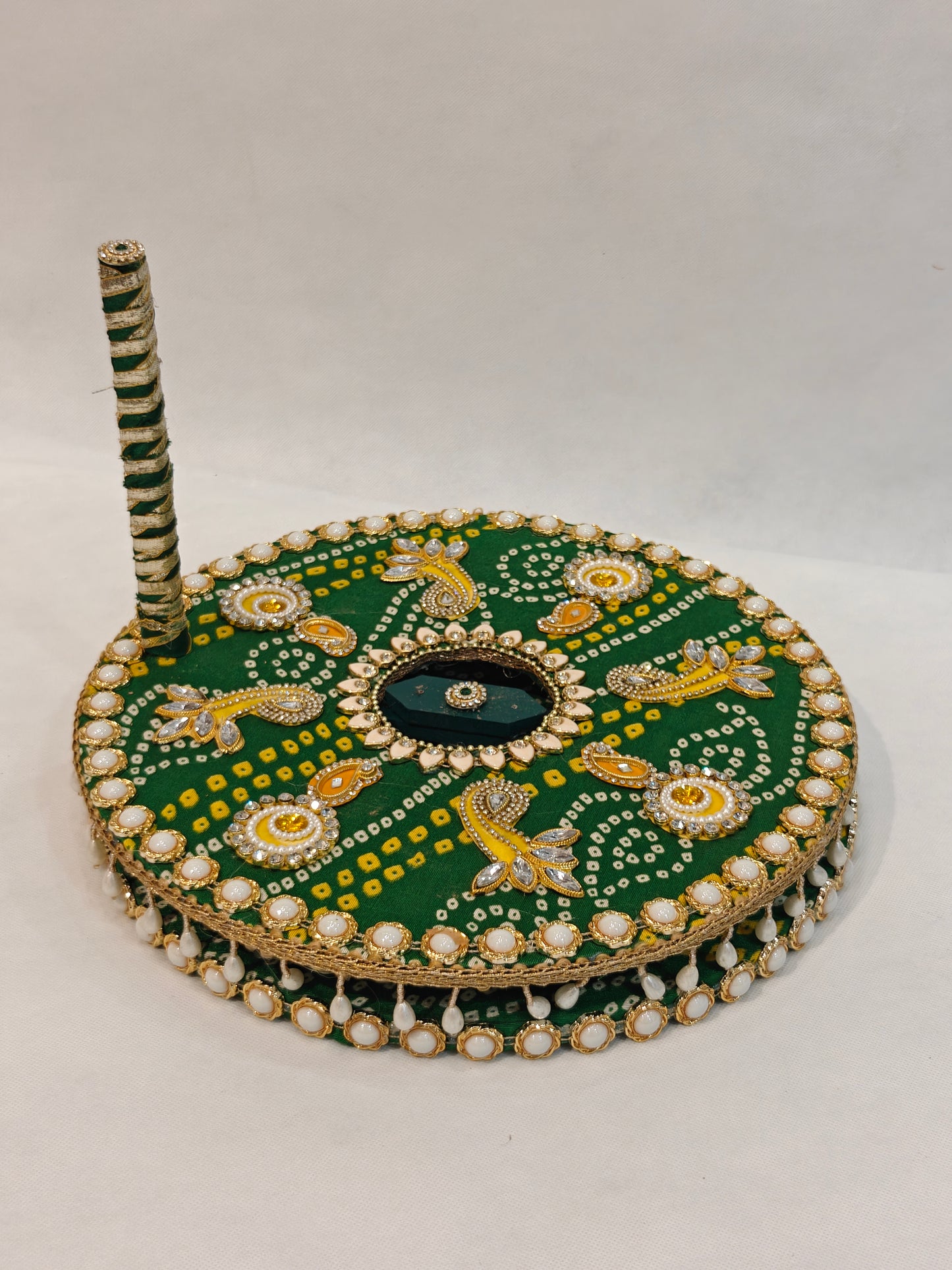 Decorative MDF chakki for Indian Baan pre-wedding ritual in green Bandhani fabric with yellow orange kundan sunburst eye motifs, gold crystal paisley, deep green hexagonal mirror centre, white kundan lotus burst, pearl cabochon border, hanging pearl drop fringe and green white ribbon wrapped handle — 12 inch diameter