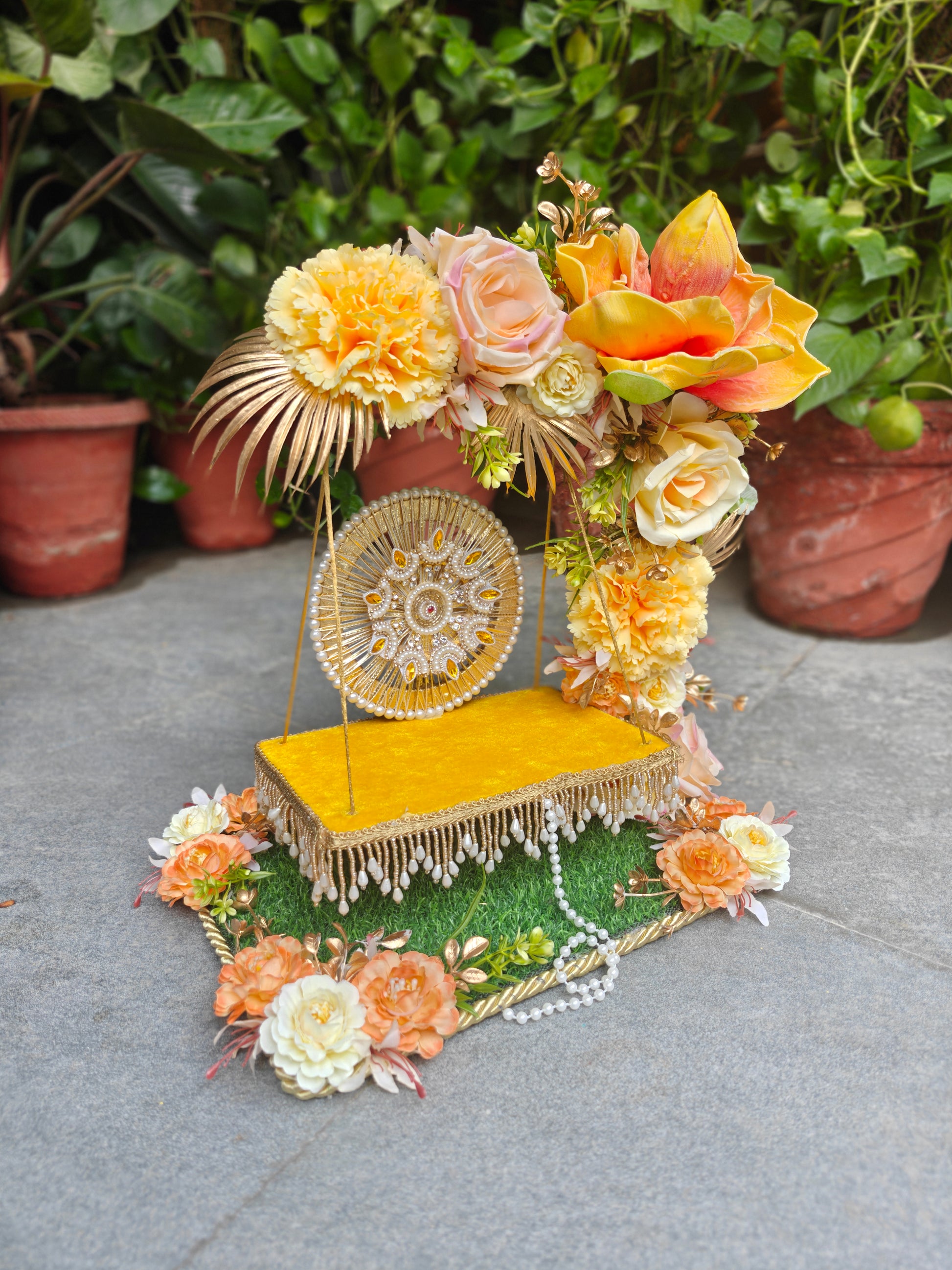 A decorative floral and ornamental display featuring a gold-framed circular mirror, yellow fabric platform, faux green grass, white pearls, and multiple orange, peach, cream, and yellow flowers, arranged on a gray stone surface with potted plants in the background.