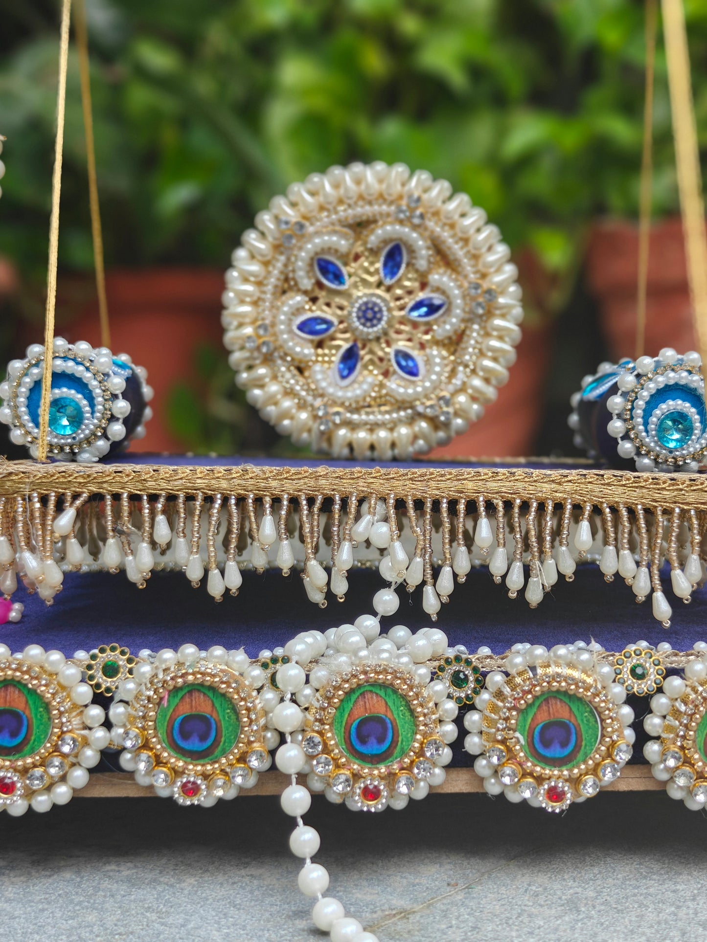 An ornate traditional Indian jewelry set featuring a large round gold and pearl necklace with blue stone accents, a matching circular pendant, and a beaded garland with peacock-eye style motifs, displayed against a soft-focus green background.