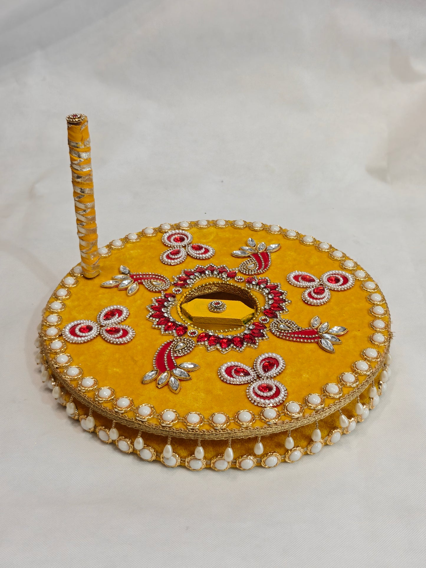 Decorative MDF chakki for Indian Baan pre-wedding ritual in mustard yellow velvet with red kundan paisley motifs, crystal rhinestones, hexagonal mirror centre inlay, white pearl edge border, hanging pearl fringe and ribbon-wrapped handle — 12 inch diameter