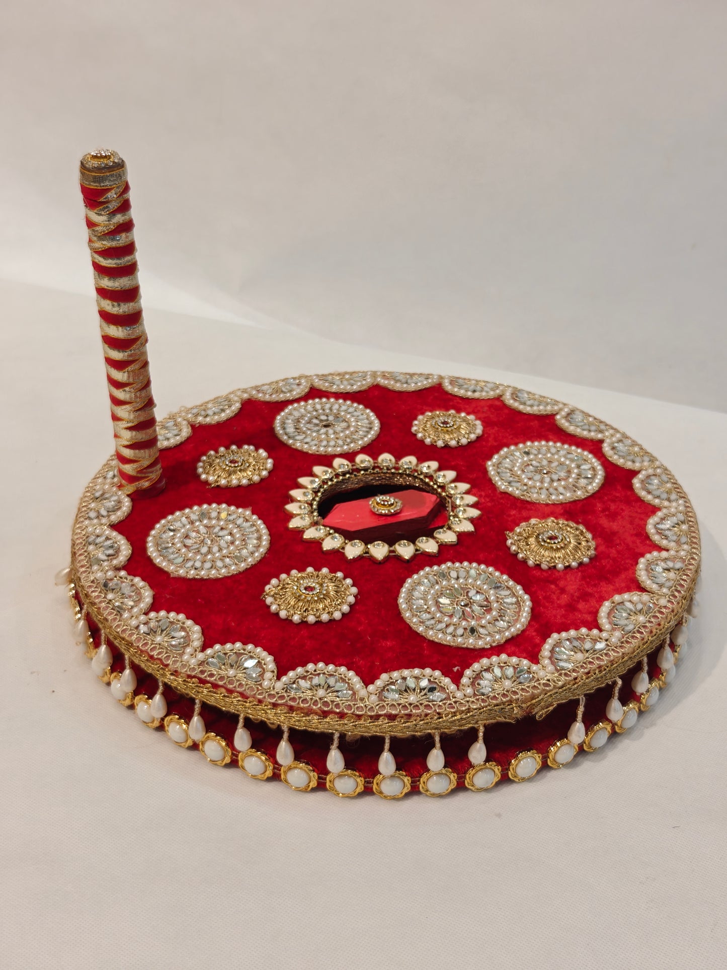 Side profile of handcrafted red velvet mirror work Baan chakki showing gold scalloped lace border, alternating hanging white pearl teardrop fringe and pearl cabochon underside detail, and red gold spiral ribbon wrapped handle with ruby kundan finial on 12 inch MDF grinding wheel for Indian pre-wedding ceremony