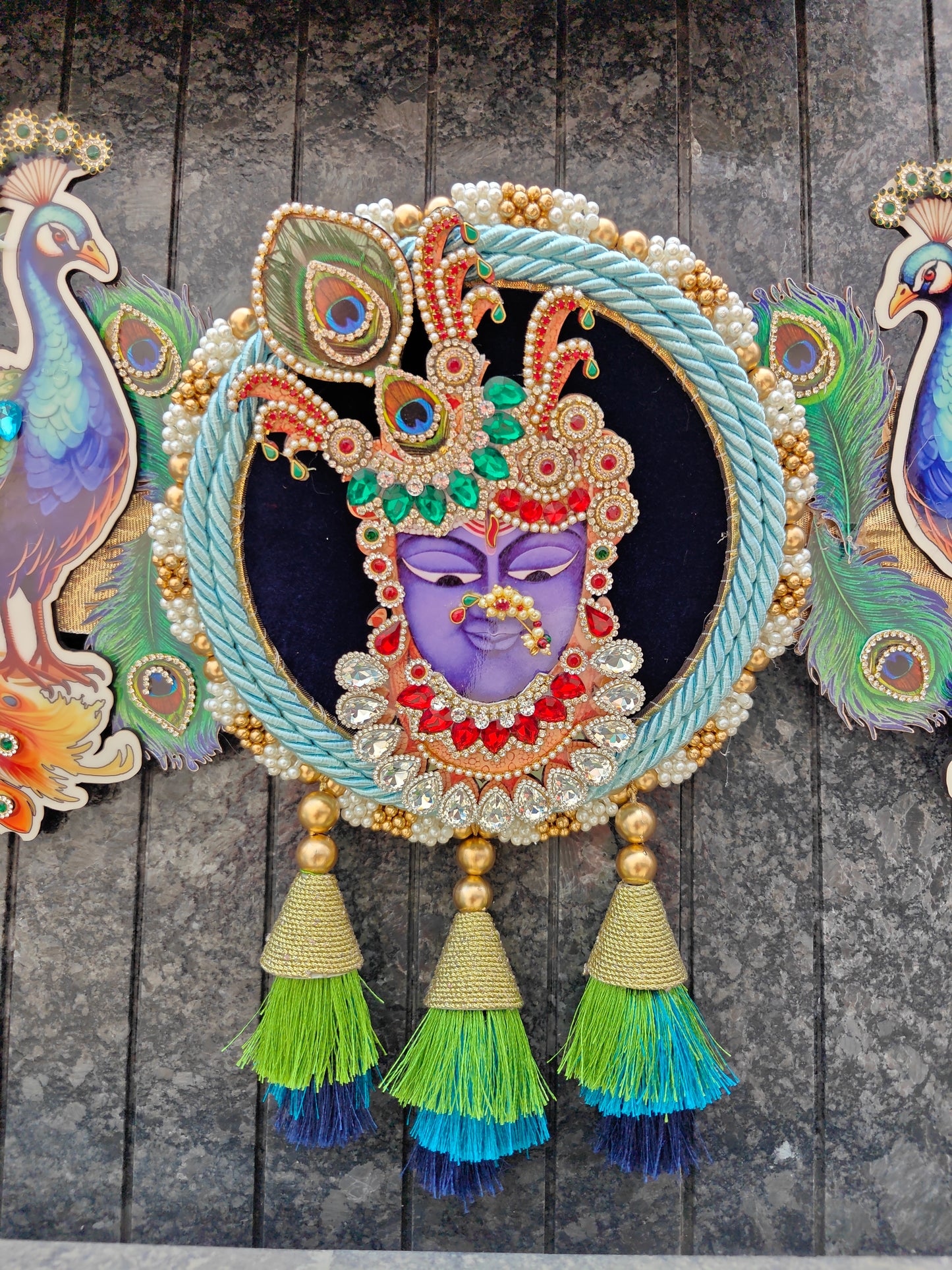 An ornate circular decorative wall hanging featuring a purple face with a jeweled headdress, surrounded by a braided light blue rope border and gold tassel accents, displayed against a dark textured wall.