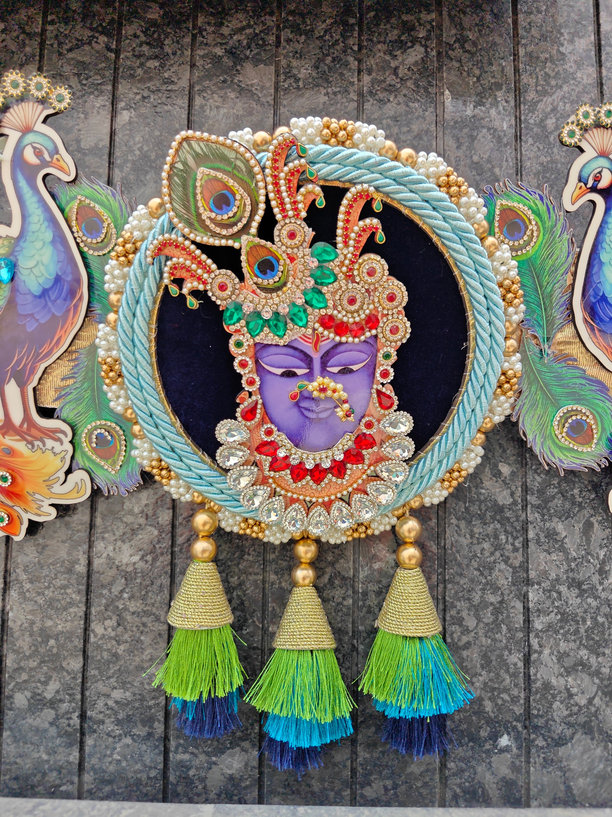An ornate circular decorative wall hanging featuring a purple face with a jeweled headdress, surrounded by a braided light blue rope border and gold tassel accents, displayed against a dark textured wall.