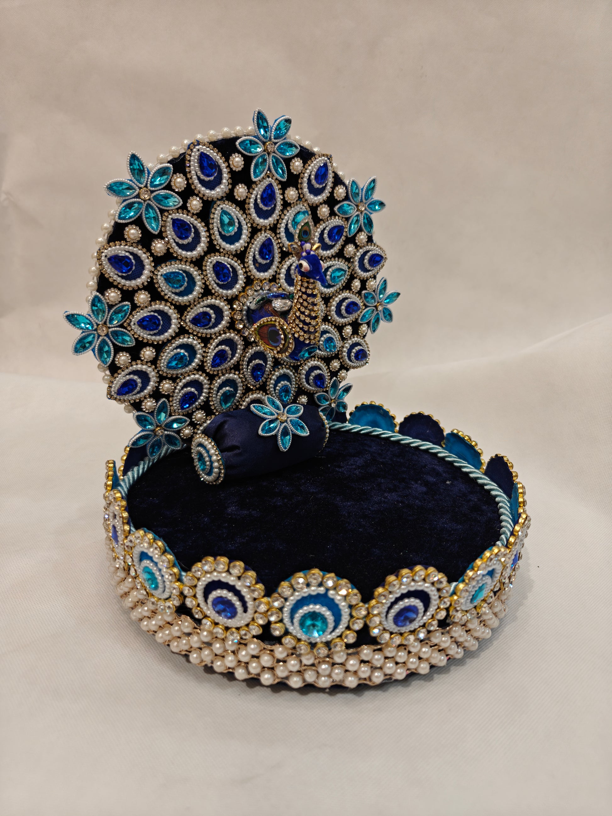 Handcrafted peacock Singhaasan for Ladoo Gopal shown at an angle — 9 inch MDF deity throne with detachable peacock backdrop, royal blue and teal gem embellishments, pearl beaded border and navy velvet base by The Wedding Station