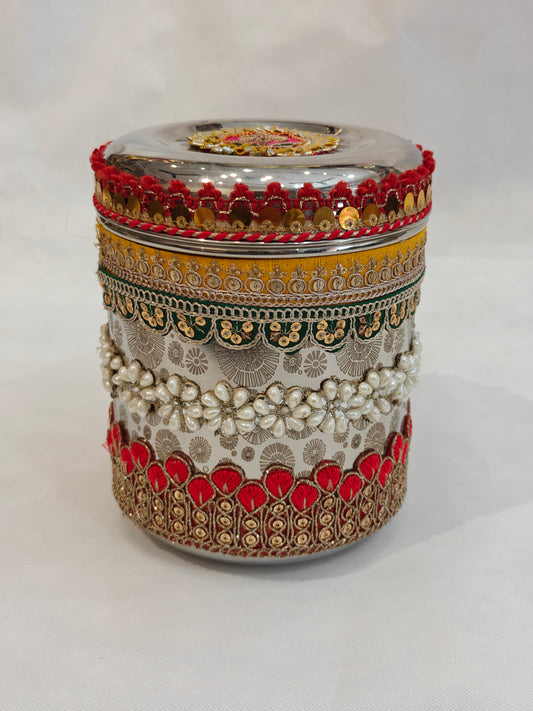 Full front view of round decorated stainless steel storage box for Indian wedding with five embellishment layers — red tassel border, mustard zari lace, green sequin trim, pearl flower cluster band and red gold teardrop patti base border, 5 inch diameter 6.5 inch height