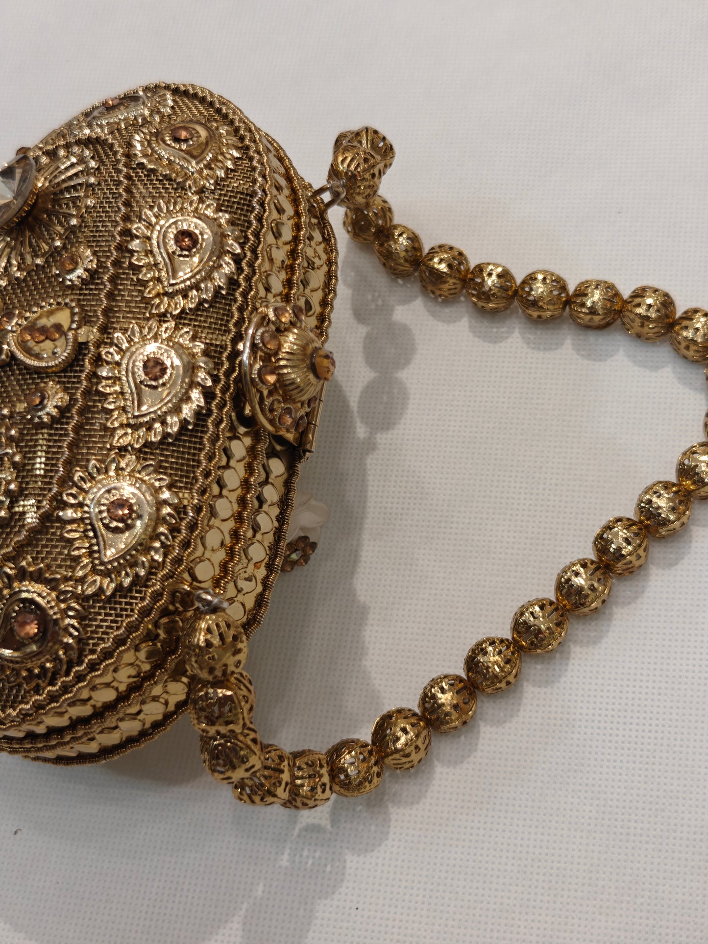 Handcrafted gold brass oval minaudière hard case clutch with champagne solitaire crystals, raised paisley filigree motifs, sunburst medallions, domed clasp finial and filigree bead top handle — bridal wedding evening bag by The Wedding Station Meerut