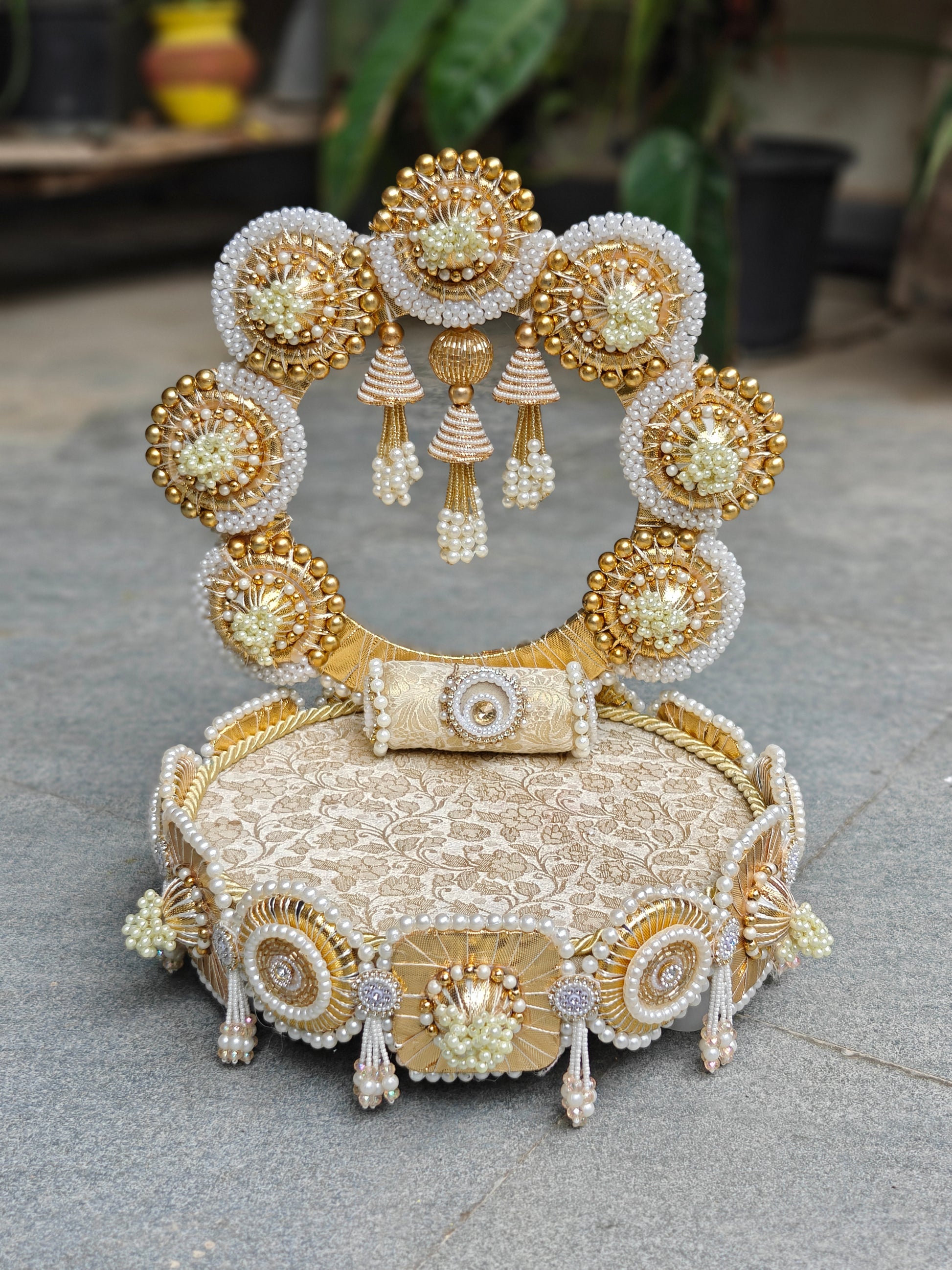 An ornate traditional Indian bridal headpiece (mukut) featuring intricate gold and white beadwork, pearl embellishments, and hanging tassels, displayed on a textured beige fabric cushion outdoors.