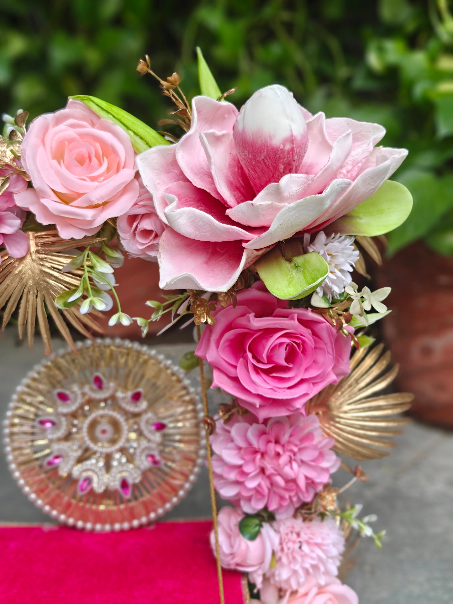 A decorative floral arrangement featuring large pink and white magnolia-style flowers, pink roses, pink dahlias, green leaves, and gold fan accents, photographed outdoors with a blurred green background and a round ornate plate in the foreground.