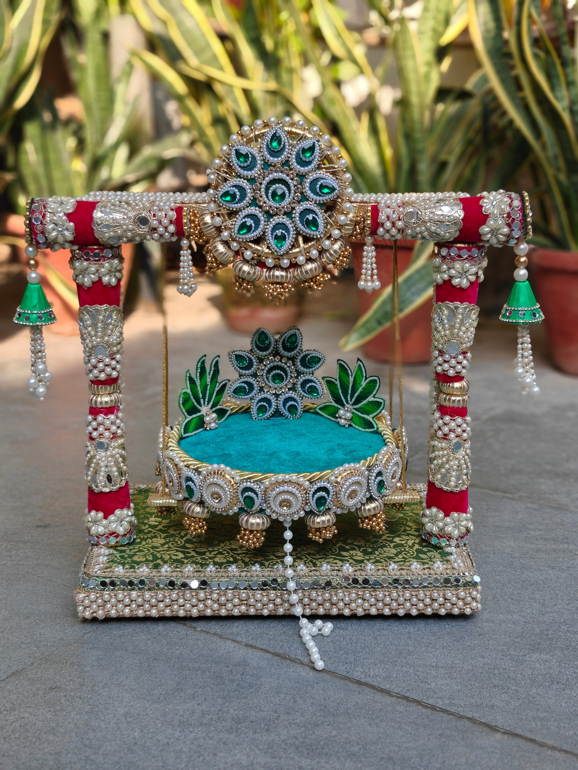 Front view of Royal Collection handcrafted Kanha jhula and singhasan in emerald green teal and crimson – emerald stone torana medallion, crimson zardozi pillars, detachable teal patra with green lotus peacock eye backrest, gold brocade MDF base with double pearl border – The Wedding Station