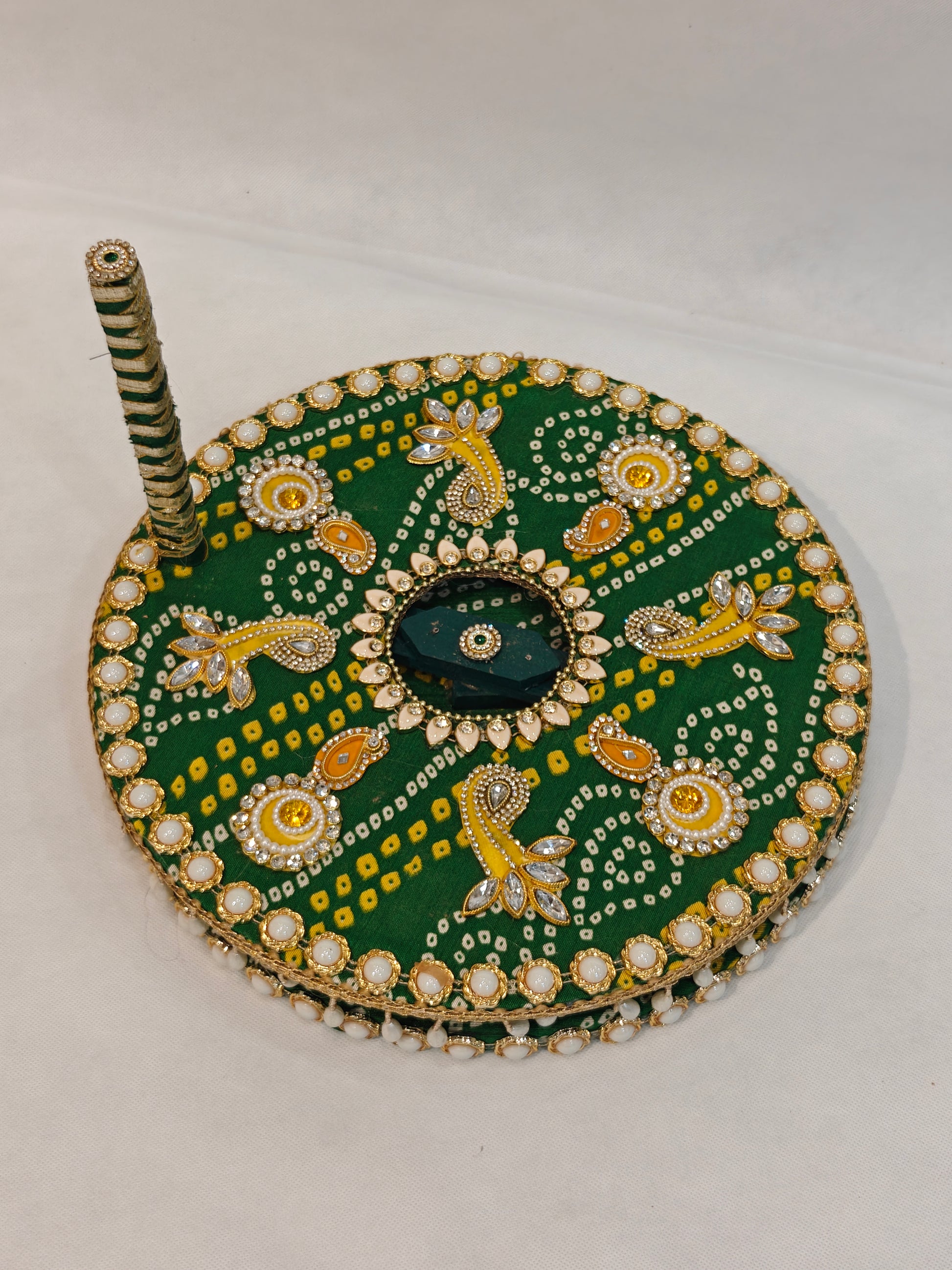 Side profile of handcrafted green Bandhani decorative Baan chakki showing white pearl cabochon edge border, hanging pearl teardrop fringe underside detail and green white spiral ribbon wrapped handle with kundan finial on 12 inch MDF grinding wheel for Indian pre-wedding ceremony