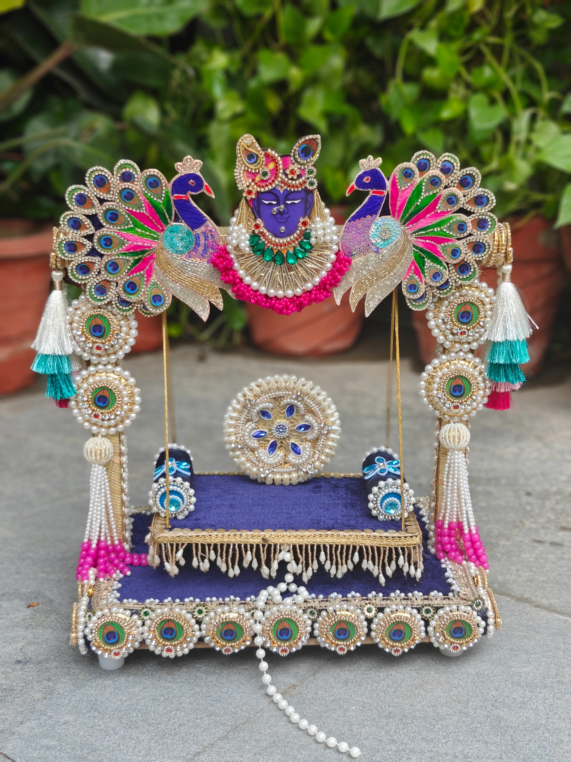 An ornate decorative miniature idol of Lord Krishna seated on a throne, embellished with gold-toned jewelry, white pearls, and vibrant blue, pink, and green accents, photographed outdoors against a blurred background of green foliage and terracotta pots.