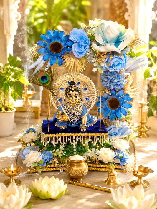 A decorative religious display featuring a seated gold-toned deity statue on a blue draped pedestal, surrounded by large blue and white flowers, green foliage, and brass oil lamps in a sunlit indoor setting.
