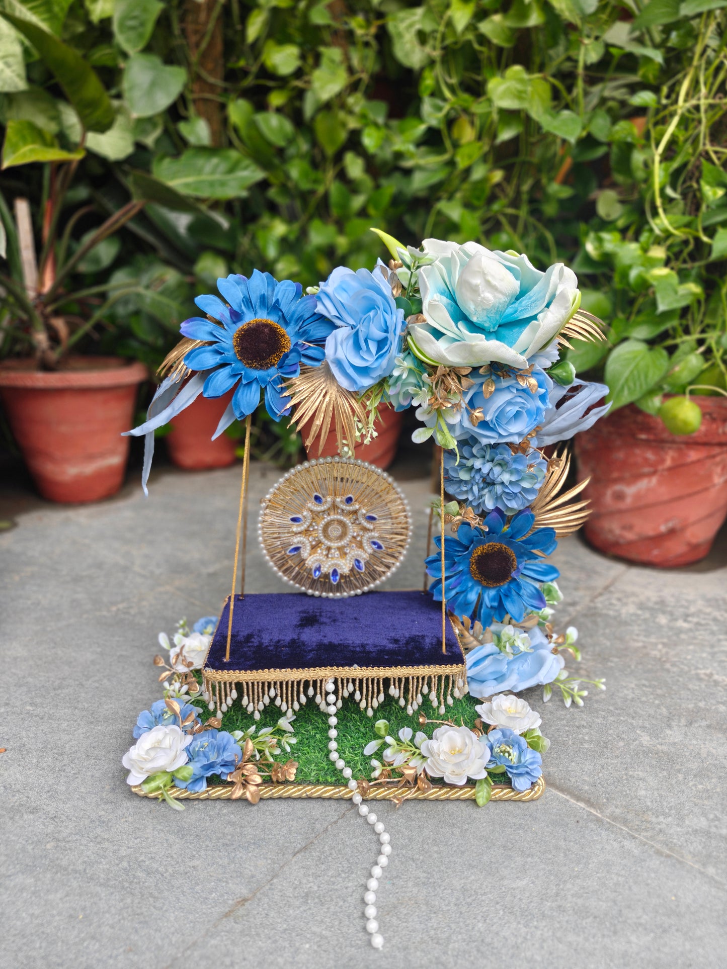 A decorative miniature stage set with a purple velvet bench, gold-framed sides, a circular gold medallion backdrop, and elaborate blue and white floral arrangements, displayed outdoors on a stone surface with potted plants in the background.