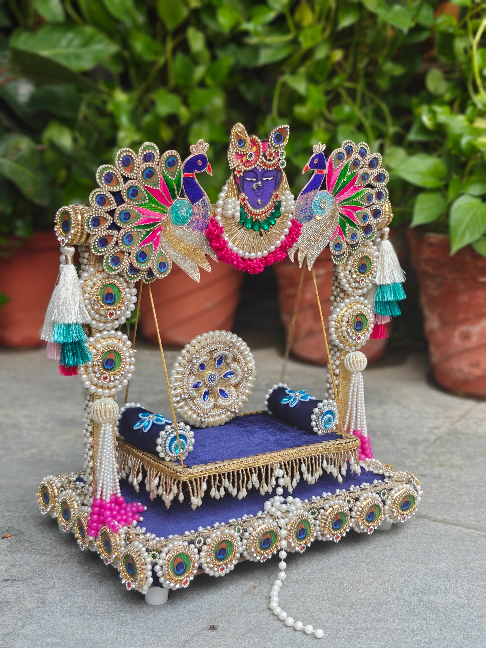 An ornate decorative peacock canopy with a purple seat, blue cushions, and intricate silver beadwork, displayed outdoors in front of green potted plants.