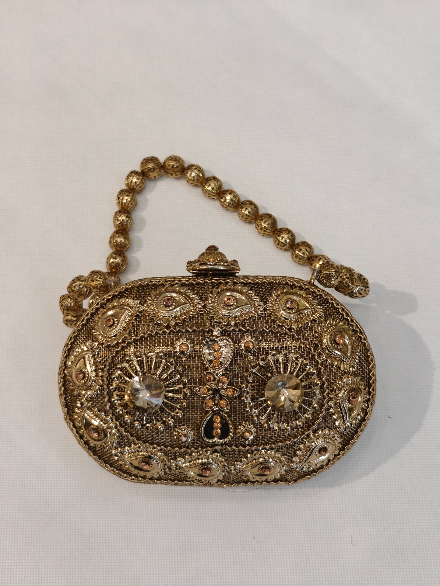 Handcrafted gold brass oval minaudière hard case clutch with champagne solitaire crystals, raised paisley filigree motifs, sunburst medallions, domed clasp finial and filigree bead top handle — bridal wedding evening bag by The Wedding Station Meerut