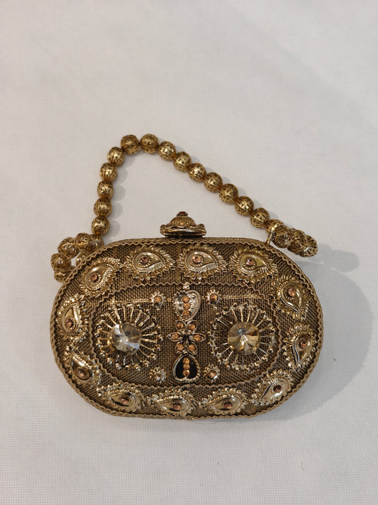 Handcrafted gold brass oval minaudière hard case clutch with champagne solitaire crystals, raised paisley filigree motifs, sunburst medallions, domed clasp finial and filigree bead top handle — bridal wedding evening bag by The Wedding Station Meerut