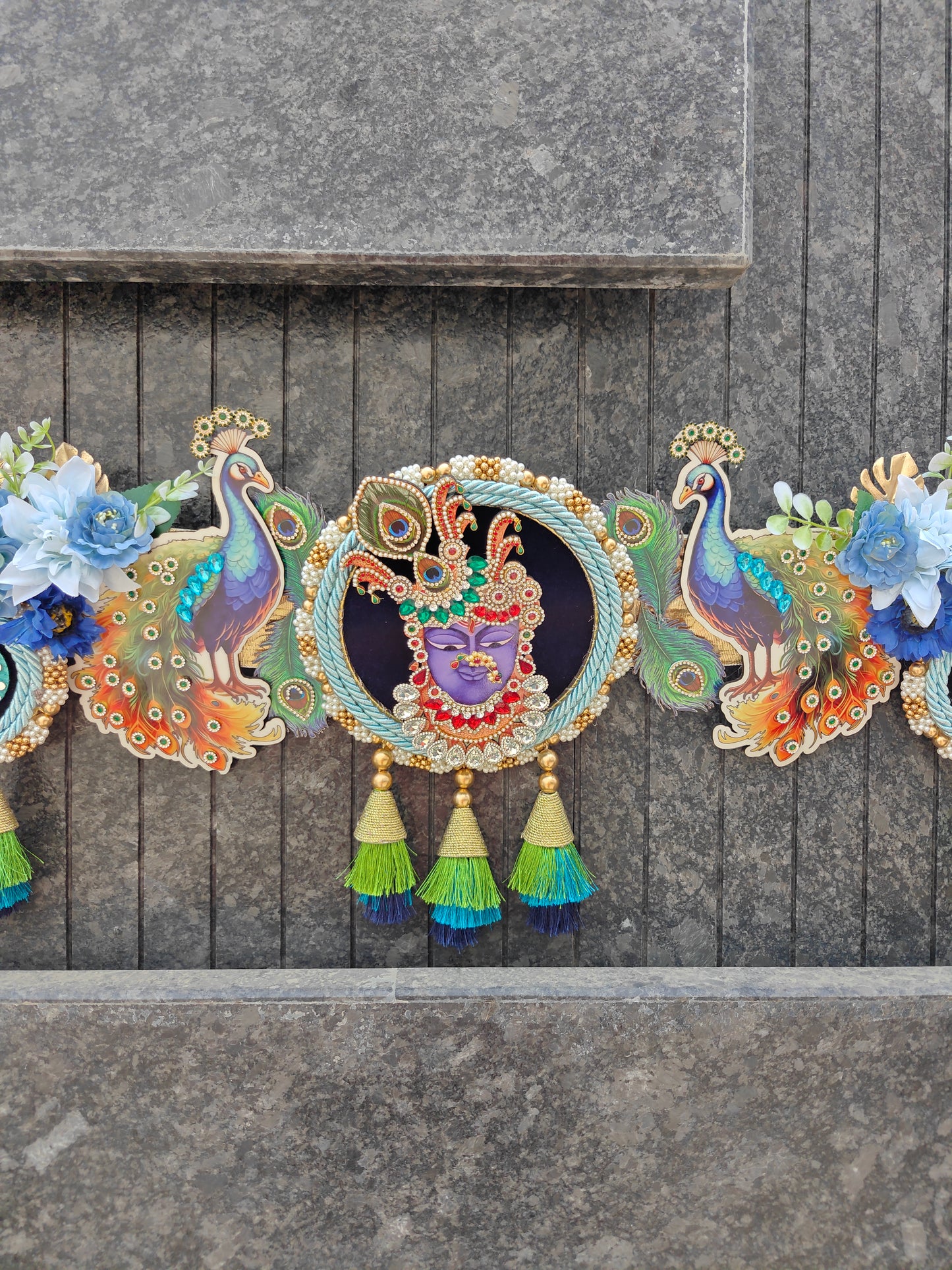 A decorative garland featuring colorful peacock and floral motifs with a central circular medallion showing a purple-faced figure, hung against a gray stone wall.