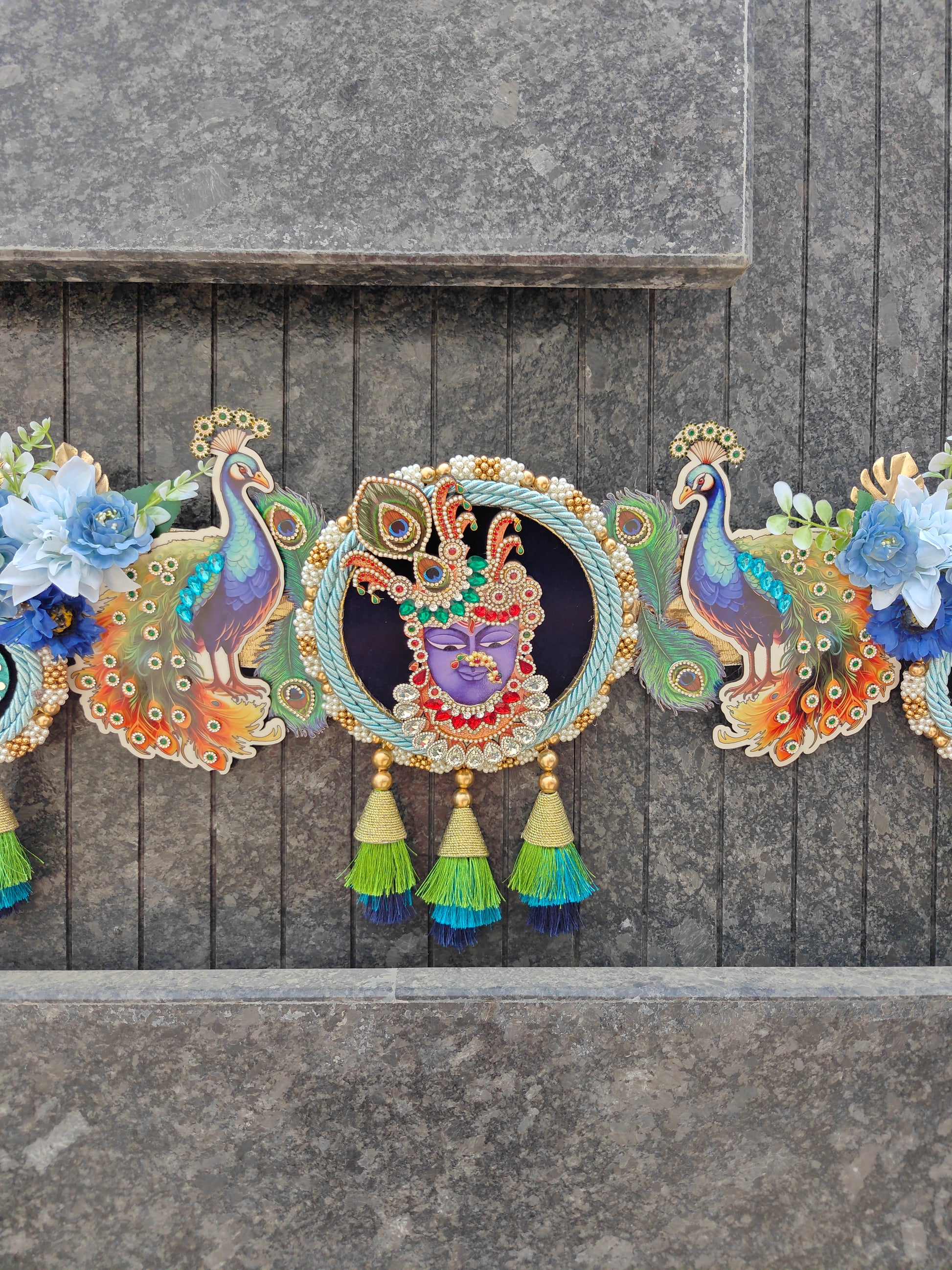 A decorative garland featuring colorful peacock and floral motifs with a central circular medallion showing a purple-faced figure, hung against a gray stone wall.