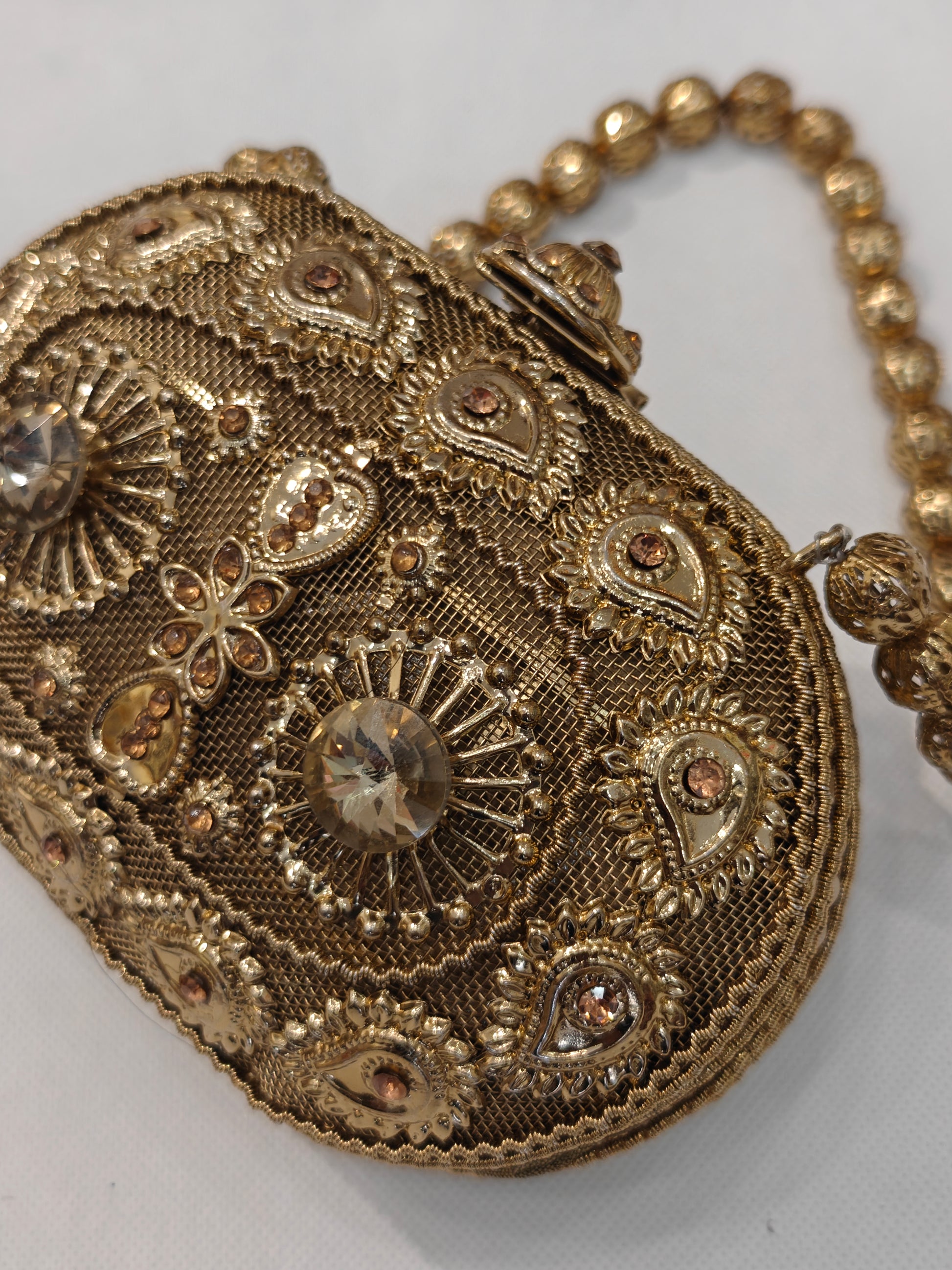 Handcrafted gold brass oval minaudière hard case clutch with champagne solitaire crystals, raised paisley filigree motifs, sunburst medallions, domed clasp finial and filigree bead top handle — bridal wedding evening bag by The Wedding Station Meerut