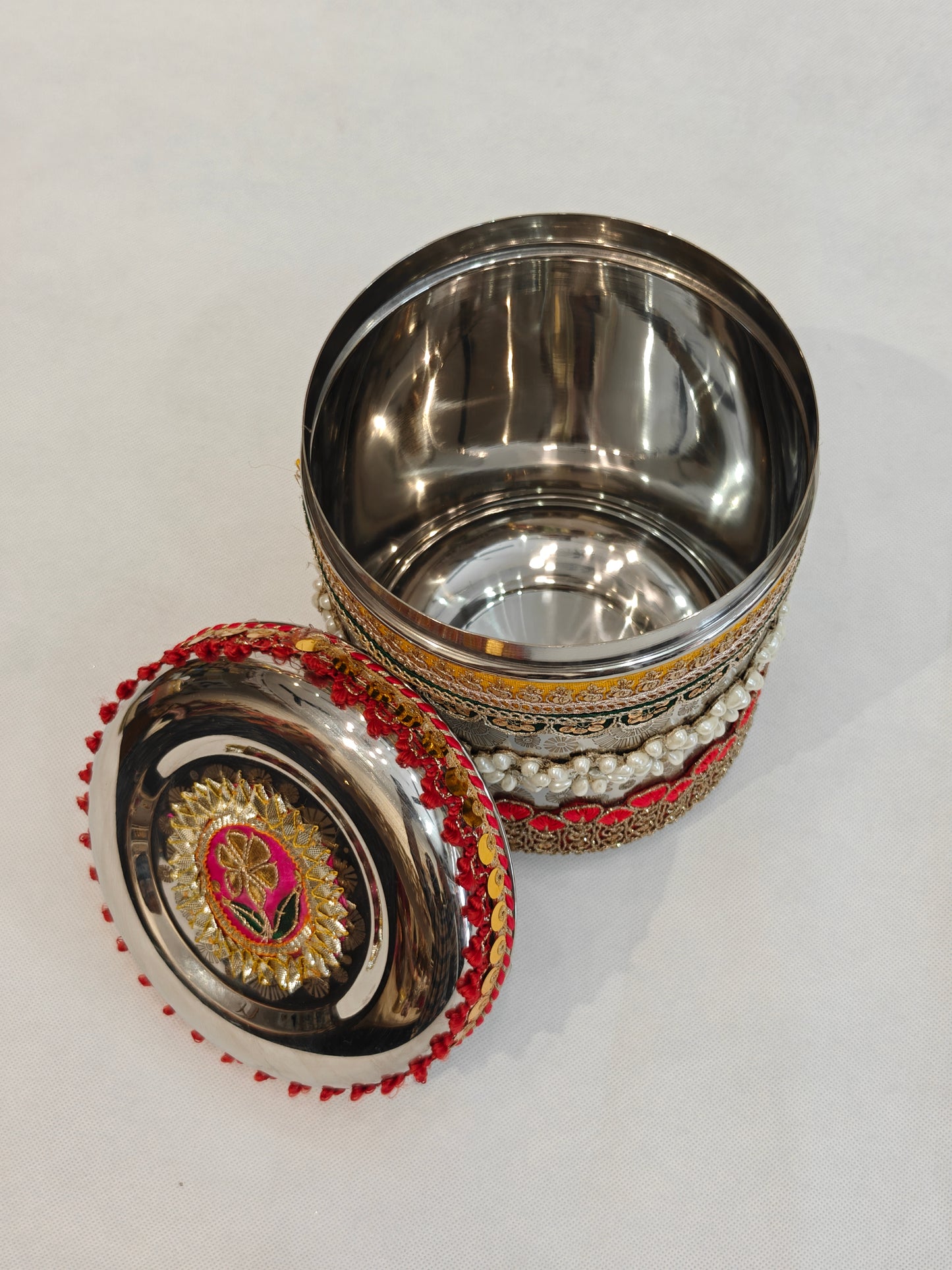 Open decorated round stainless steel storage box showing mirror-finish interior and gota patti zardozi floral medallion lid with pink, gold and green motif, red tassel rim, and multi-layer pearl and zari embellished body for Indian wedding trousseau