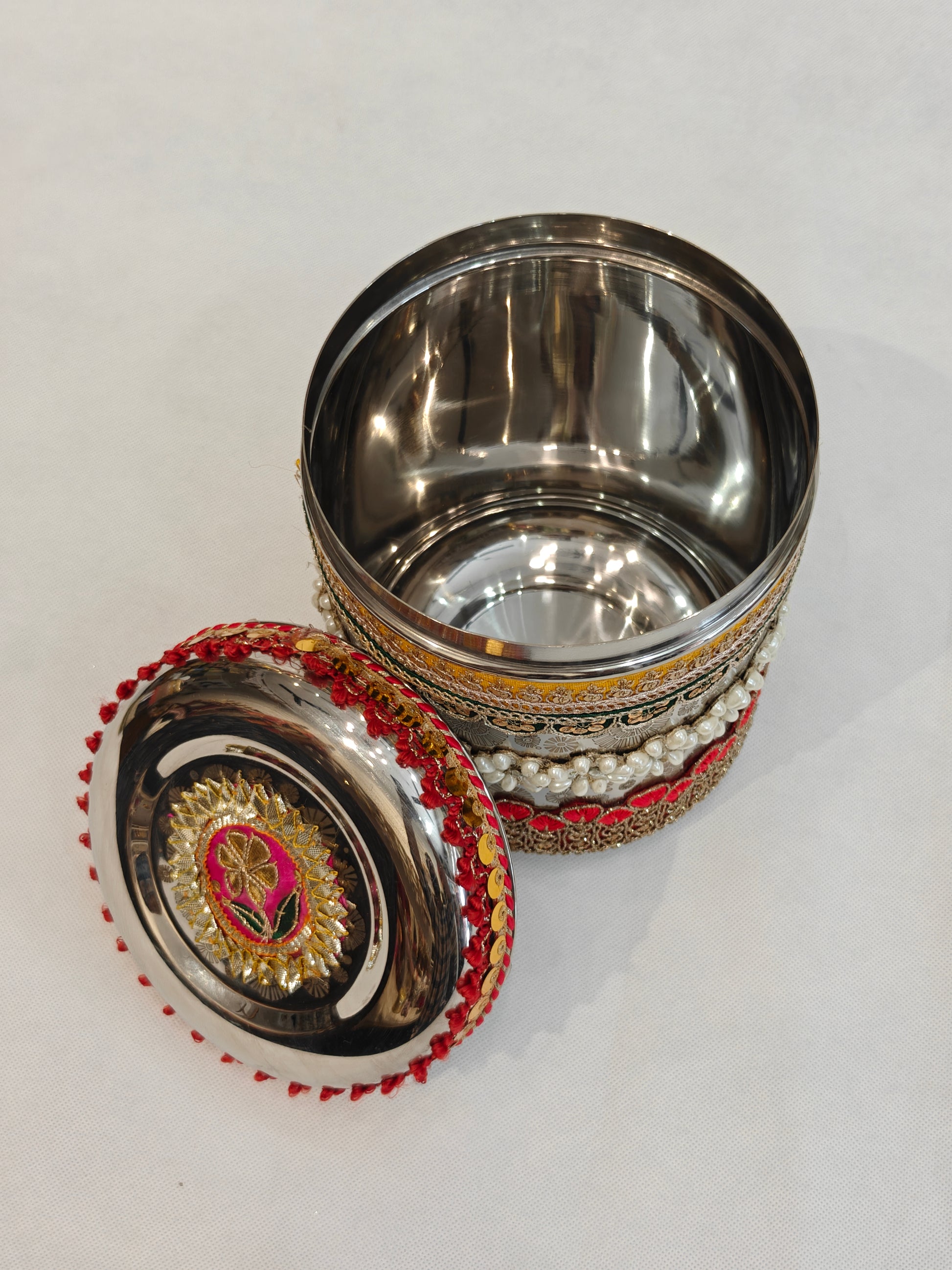 Open decorated round stainless steel storage box showing mirror-finish interior and gota patti zardozi floral medallion lid with pink, gold and green motif, red tassel rim, and multi-layer pearl and zari embellished body for Indian wedding trousseau