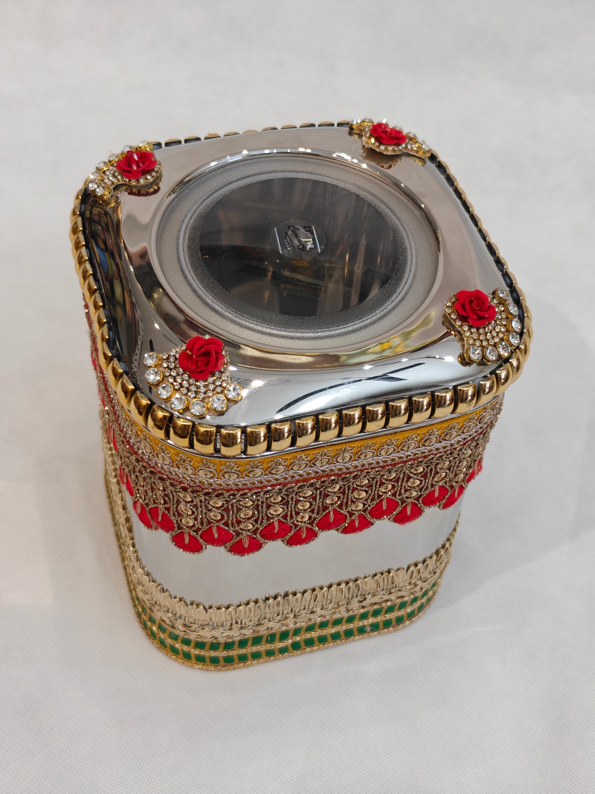 Top view of decorated stainless steel square wedding box showing four red rose kundan corner medallions, gold ridged rim and circular transparent acrylic window on lid for Indian trousseau and gifting
